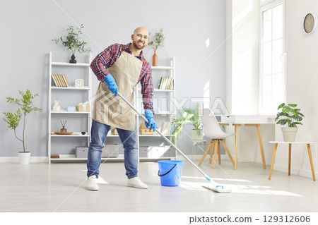 Portrait of smiling man looking cheerful at camera holding mop in hand and mopping house or office. Portrait of smiling man looking cheerful at camera holding mop in hand and mopping house or office. 129312606
