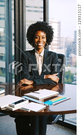 Professional businesswoman sitting confidently with crossed arms at desk, large windows revealing city skyline, embodying corporate success and leadership 129313117