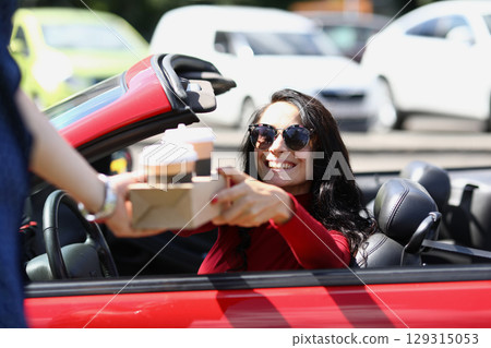 Excitement brews as a woman enjoys her coffee in a convertible on a sunny day at a bustling cafe drive-thru location 129315053