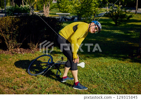 Cyclist holding injured knee while standing beside fallen bicycle 129315845