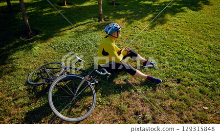Cyclist resting on grass beside bicycle in shaded park area 129315848