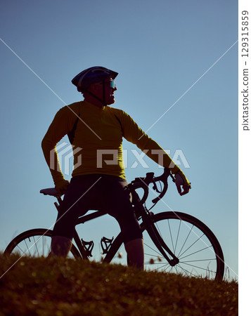 Older cyclist straddling bike and gazing sideways on grassy hilltop at dusk 129315859