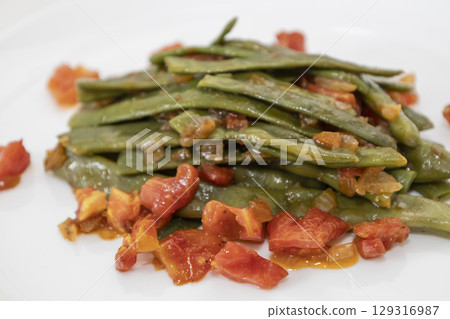 Fried green beans with tomato and onion pieces. Tasty meal with french bean on dinner or lunch on the white plate Fried green beans with tomato and onion pieces. Tasty meal with french bean on dinner or lunch on the white plate 129316987