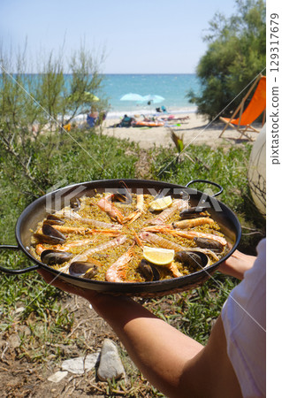 a person holds a seafood paella near the beach 129317679