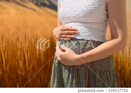 Pregnant Woman in Wheat Field. Close-up of a pregnant woman gently holding her belly while standing in a golden wheat field. Pregnant Woman in Wheat Field. Close-up of a pregnant woman gently holding her belly while standing in a golden wheat field. 129317750