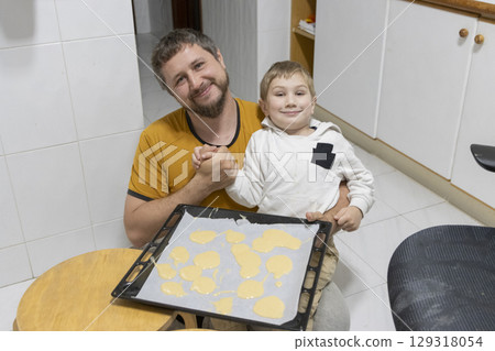 Father and son holding hands and showing baking pan with uncooked cookies in kitchen 129318054