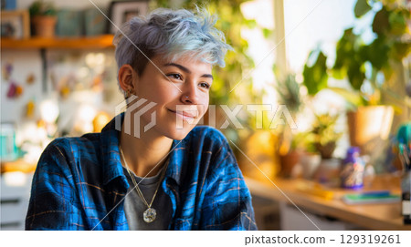 Young non-binary person with light blue hair and a joyful smile enjoys a sunny day indoors, surrounded by plants and a creative workspace, while wearing a plaid shirt 129319261