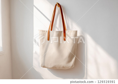 Stylish beige canvas tote bag for mockup hanging against a bright wall illuminated by soft afternoon light 129319391