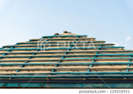 Wooden framework of a roof under construction, featuring blue-green planks against a clear sky, indicating ongoing building activity Wooden framework of a roof under construction, featuring blue-green planks against a clear sky, indicating ongoing building activity 129319551