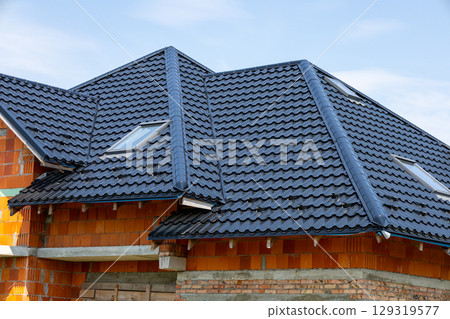 A modern residential construction showcases a detailed roof design with dark tiles and skylights, set against a clear blue sky 129319577