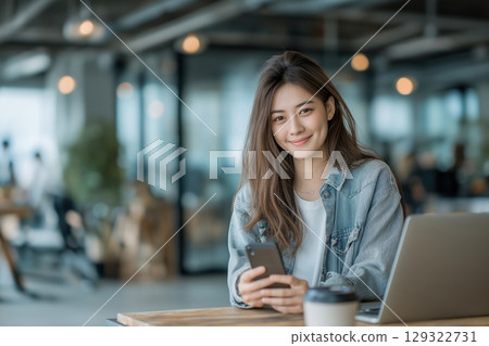 Young asian woman using mobile phone and laptop in a modern co-working space smiling charmingly 129322731