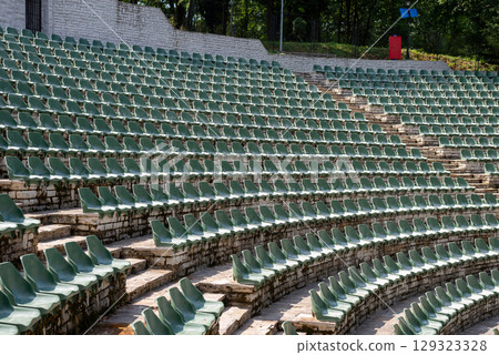 Large, open-air stone amphitheater is filled with numerous rows of green plastic seats arranged in a semi-circular pattern 129323328