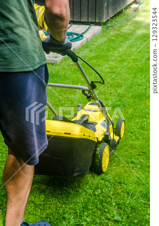 Rear view of man in shorts cutting grass with electric mower in backyard. Summer gardening and lawn care concept. High quality photo Rear view of man in shorts cutting grass with electric mower in backyard. Summer gardening and lawn care concept. High quality photo 129323544
