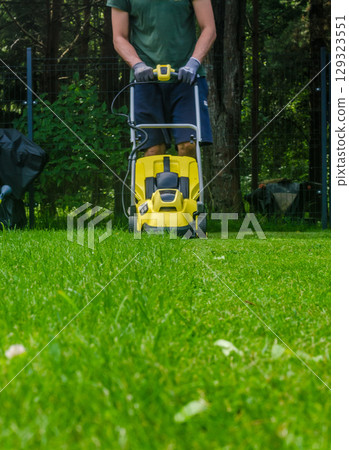 A man in shorts and gloves uses a yellow electric lawn mower on a green lawn during summer. High quality photo A man in shorts and gloves uses a yellow electric lawn mower on a green lawn during summer. High quality photo 129323551