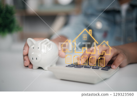 Mortgage, home loan planning, and real estate investment. Person calculating home payments using calculator with piggy bank and digital overlay of house icons and documents. Mortgage, home loan planning, and real estate investment. Person calculating home payments using calculator with piggy bank and digital overlay of house icons and documents. 129323560