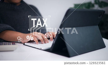 Online tax calculation, financial planning, and digital accounting service. Person using tablet and keyboard with digital overlay of TAX, calculator, percentage, and money icons. 129323561