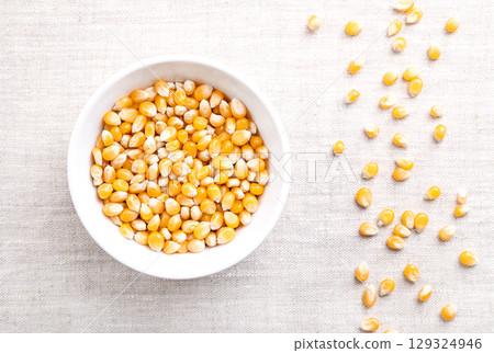 Popcorn kernels, in a white bowl on linen. Seeds of Zea mays variety everta, rounded at the top, which expand and puffs up, when heated. Snack food, sometimes sold as pearls, or as old maids. 129324946