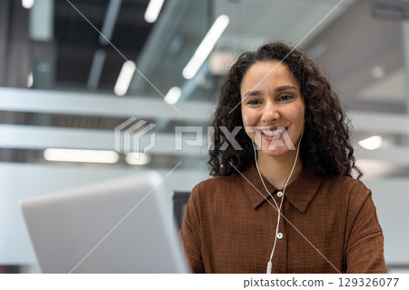 A cheerful professional woman engaging in remote work, using a laptop and earphones in an office setting. The modern environment highlights a productive and joyful workspace. A cheerful professional woman engaging in remote work, using a laptop and earphones in an office setting. The modern environment highlights a productive and joyful workspace. 129326077