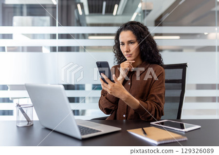 Businesswoman using a smartphone while working on a laptop in an office setting, surrounded by modern design and professional tools, reflecting focus, productivity, and digital communication Businesswoman using a smartphone while working on a laptop in an office setting, surrounded by modern design and professional tools, reflecting focus, productivity, and digital communication 129326089