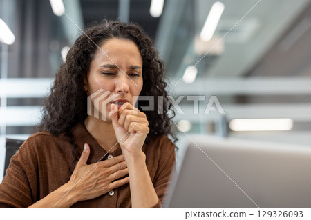 A woman coughing indoors while working in an office setting, depicting sickness or discomfort in a professional environment. Image conveys health concerns, ailment, or allergy in workplace scenarios. 129326093