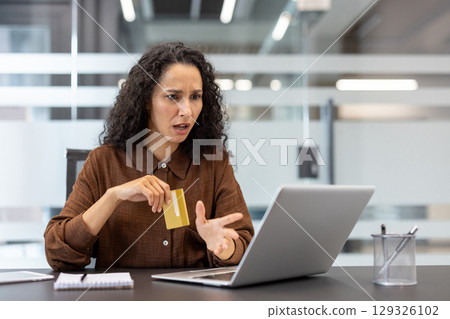 A businesswoman sitting at a desk, expressing frustration holding a credit card and using laptop. The image conveys concepts of online challenges, ecommerce issues, or digital financial difficulties. A businesswoman sitting at a desk, expressing frustration holding a credit card and using laptop. The image conveys concepts of online challenges, ecommerce issues, or digital financial difficulties. 129326102