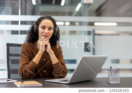 Professional woman sitting at desk, wearing headphones, working on a laptop in a modern office setting. The atmosphere reflects a productive environment, expressing focus, confidence, and dedication. Professional woman sitting at desk, wearing headphones, working on a laptop in a modern office setting. The atmosphere reflects a productive environment, expressing focus, confidence, and dedication. 129326103