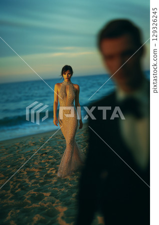 A woman in a gold dress is standing on the beach with a man in a black suit behi 129326245