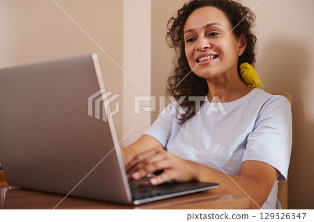 Smiling Woman Working on Laptop with a Yellow Pet Bird Perched on Her Shoulder Smiling Woman Working on Laptop with a Yellow Pet Bird Perched on Her Shoulder 129326347