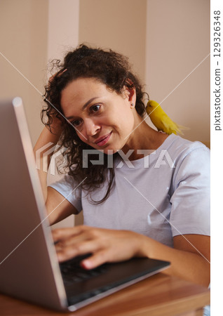 Woman Working on Laptop at Home with Yellow Bird on Her Shoulder 129326348