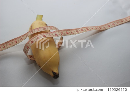 A short yellow banana wrapped with a measuring tape, placed on a clean white background. This image highlights both the length and circumference of the fruit. A short yellow banana wrapped with a measuring tape, placed on a clean white background. This image highlights both the length and circumference of the fruit. 129326350