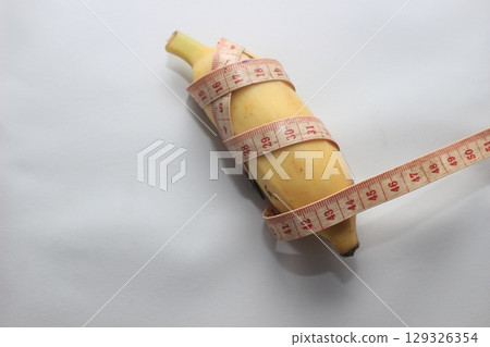 A short yellow banana wrapped with a measuring tape, placed on a clean white background. This image highlights both the length and circumference of the fruit. 129326354