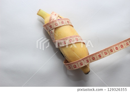 A short yellow banana wrapped with a measuring tape, placed on a clean white background. This image highlights both the length and circumference of the fruit. 129326355