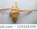 A short yellow banana wrapped with a measuring tape, placed on a clean white background. This image highlights both the length and circumference of the fruit. 129326356