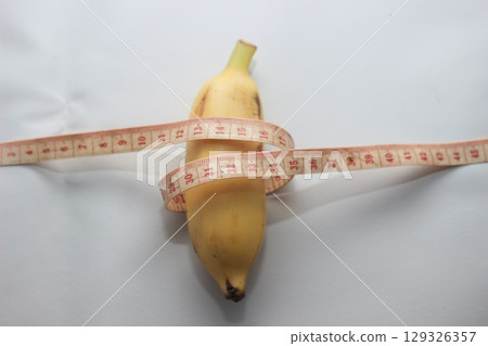 A short yellow banana wrapped with a measuring tape, placed on a clean white background. This image highlights both the length and circumference of the fruit. 129326357