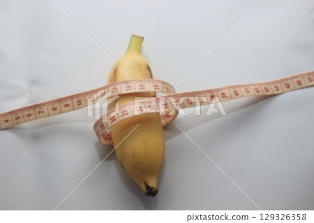 A short yellow banana wrapped with a measuring tape, placed on a clean white background. This image highlights both the length and circumference of the fruit. 129326358