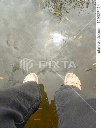 Person's legs in sandals with a tranquil and moody feeling dangling over water, representing relaxation and contemplation against a water reflection background 129327414