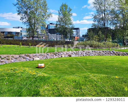 Automated lawn mower navigating a well-maintained grassy area, surrounded by landscaped flowers and rocks. Robot lawnmower Automated lawn mower navigating a well-maintained grassy area, surrounded by landscaped flowers and rocks. Robot lawnmower 129328011
