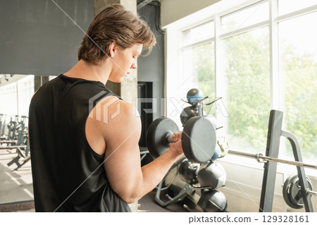 Muscular young caucasian man lifting dumbbell in gym setting. Weightlifting exercises, physical fitness. Work out. 129328161