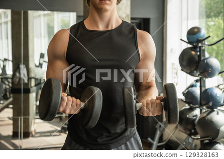 Close up of muscular arm lifting dumbbells in gym setting. Weightlifting exercises, physical fitness. Bodybuilder lifting dumbbells, biceps exercise in gym. 129328163