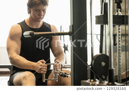 Young sporty caucasian man wearing blank sleeveless tank top exercising on low rowing machine at gym. 129328168