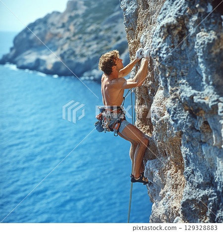 Man climbs steep cliff above a blue sea, using a rope and gear. Concept of adventure and physical challenge. For sports content. 129328883
