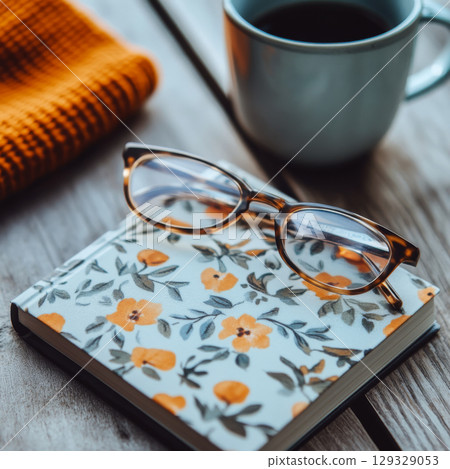 Glasses sit on a floral-patterned notebook by a mug and knitted cloth on a wooden table. Concept of cozy and creative workspace. For editorial or article photo use. 129329053