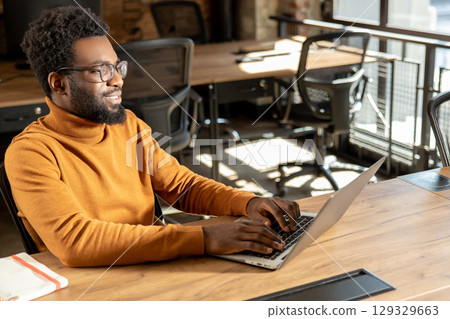 Bearded man in glasses typing on laptop working online from stylish industrial-style office Bearded man in glasses typing on laptop working online from stylish industrial-style office 129329663