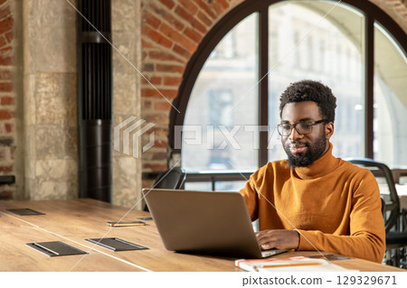 Bearded man in glasses typing on laptop working online from stylish industrial-style office 129329671