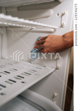 Vertical cropped shot of unrecognizable person defrosting and cleaning interior of refrigerator using blue rag to wipe away ice and grime buildup, ensuring fresh and sanitary food storage environment. 129330273
