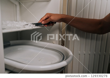 Person carefully defrosting their refrigerator, using knife to scrape off ice buildup and collecting ice chips in basin placed inside appliance for easy cleanup. Concept of domestic work, housekeeping 129330278