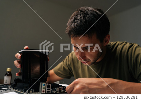 Close-up portrait of skilled computer technician male carefully installing CPU cooler onto motherboard, performing maintenance and upgrade service on desktop computer in dark workshop environment. Close-up portrait of skilled computer technician male carefully installing CPU cooler onto motherboard, performing maintenance and upgrade service on desktop computer in dark workshop environment. 129330281