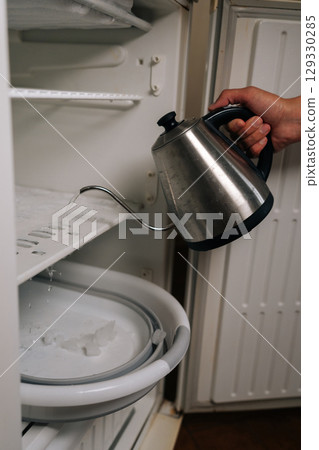 Housekeeper pouring hot water from kettle into refrigerator to melt ice and defrost, collecting melted ice in basin placed at bottom, close-up. Concept of need for defrosting due to excessive buildup. 129330285
