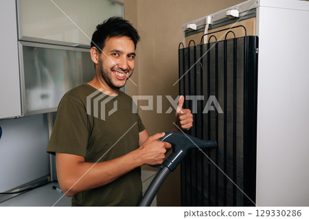 Friendly cleaning service employee vacuuming coils of refrigerator using vacuum cleaner, giving thumbs up gesture to indicate job well done, smiling looking at camera. Concept of hygiene, cleanliness. Friendly cleaning service employee vacuuming coils of refrigerator using vacuum cleaner, giving thumbs up gesture to indicate job well done, smiling looking at camera. Concept of hygiene, cleanliness. 129330286