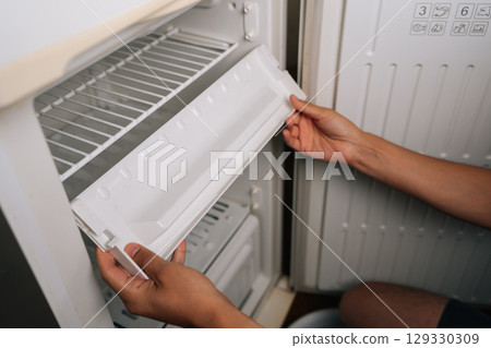 Man preparing to removing ice tray from refrigerator during defrosting, suggesting thorough cleaning process for household appliance maintenance. Concept of need for defrosting to excessive buildup 129330309
